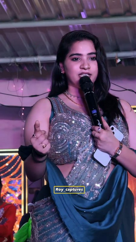 Young woman in a sparkly blue-silver sari singing into a microphone on a stage with purple lighting.