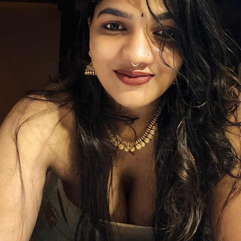 Close-up selfie of smiling South Asian woman wearing traditional gold jewelry, warm soft indoor l...