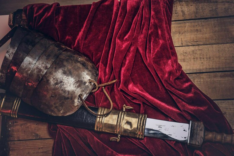 Rusty medieval sword with brass hilt and worn metal gauntlet on red velvet over a wooden floor.