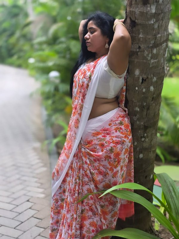Woman in a floral saree leaning against a tree in a lush outdoor garden.