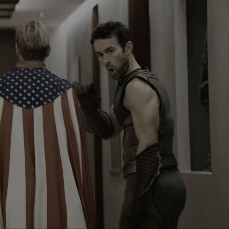 Muscular man in armor glances over his shoulder in a dim hallway, with a person in an American fl...