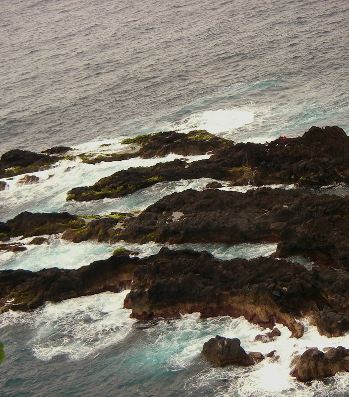 High-angle view of turquoise ocean waves crashing on mossy dark volcanic coast, with two tiny hik...
