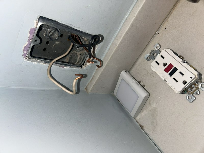 Exposed electrical box with loose wires, a detached wall plate, and a nearby outlet on a light gr...