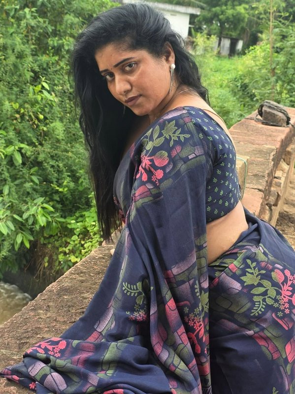 South Indian woman in a navy floral saree looks over her shoulder seated on a rural stone bridge ...