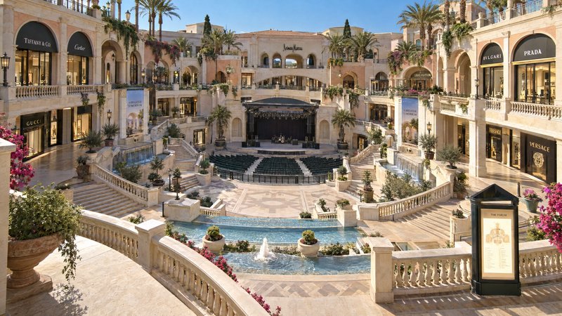 Ornate open-air luxury shopping mall courtyard with tiered fountains, designer boutiques, and a c...