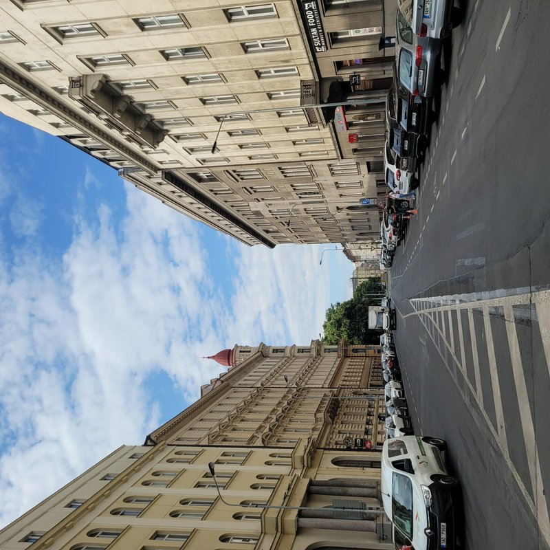 Tilted daytime view of a Central European city street lined with historic stone buildings, parked cars, and cloudy blue sky.