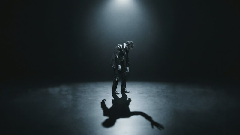 Man in a suit under a single spotlight on stage, bent over with a dramatic shadow cast on the floor.