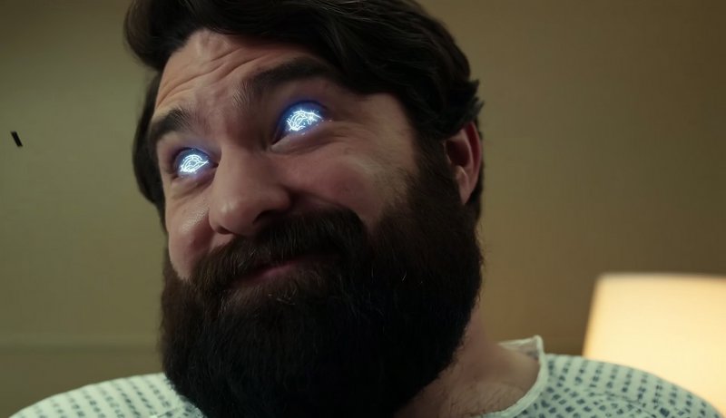 Bearded man in a hospital gown with glowing blue eyes looking upward.