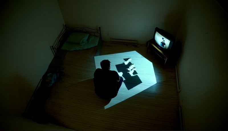 High-angle shot of a silhouetted person in a dark bedroom, sitting before a floor projection and ...