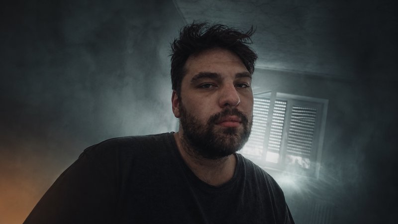 Low angle portrait of a bearded man with messy dark hair, standing in a hazy dim indoor room near...