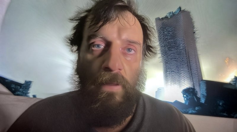 Close-up selfie of a disheveled bearded man with a startled expression, with a crumbling apocalyp...