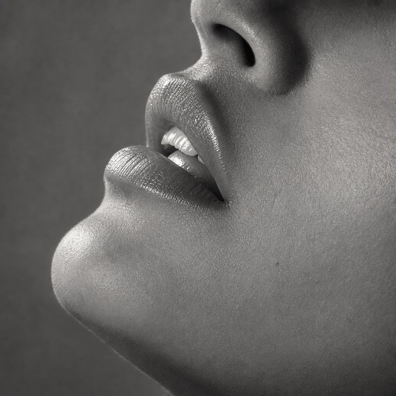 Monochrome close-up studio shot of a person's lower face, showing glossy parted lips, smooth skin...
