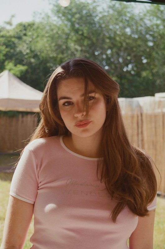 Young woman outdoors in a pink t-shirt with long brown hair, soft sunlight and a wooden fence bac...