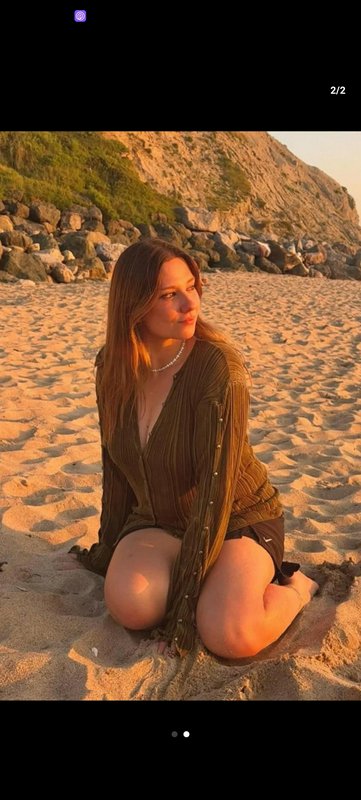 Woman sitting on a sandy beach at sunset with a rocky coastline in the background.