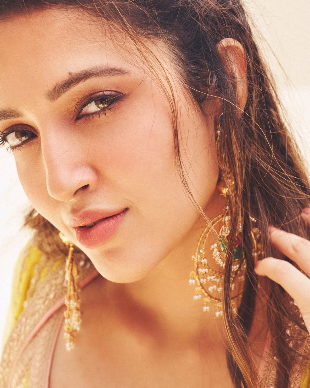 Close-up portrait of a woman with soft makeup, warm tones, and gold jewelry.