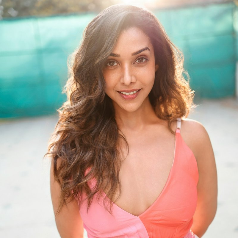 Portrait of a smiling woman with long wavy brown hair in a pink sleeveless dress outdoors.