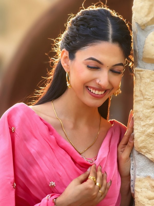 Smiling Indian woman in a pink traditional outfit with gold jewelry beside a stone wall.