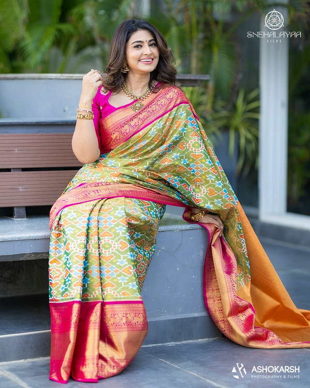 Smiling woman modeling a vibrant green and pink Patola silk saree for Snehalaayaa Silks, seated o...