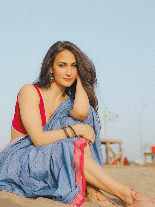 Actress Elli AvrRam sits on a sandy beach in a blue polka dot saree and red blouse, with henna on...