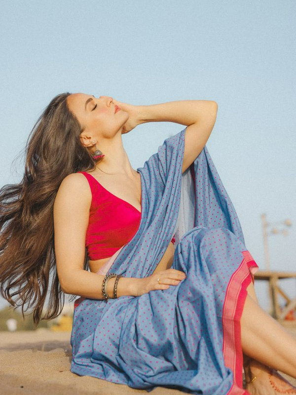 Woman in blue polka dot saree with red blouse sits on a sandy beach, eyes closed, wind blowing th...