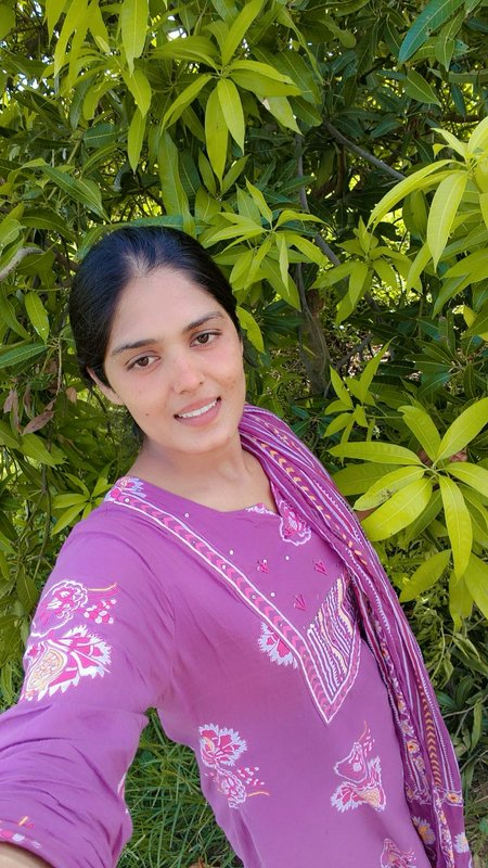 Smiling South Asian woman in embroidered purple traditional wear takes a selfie beside lush green...