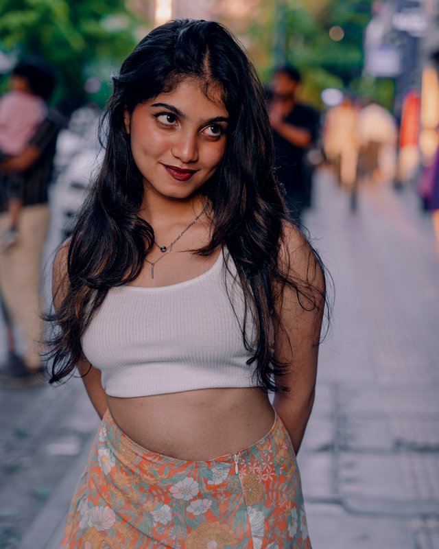 Young South Asian woman with long wavy dark hair smiles softly, standing on a dusk city sidewalk ...