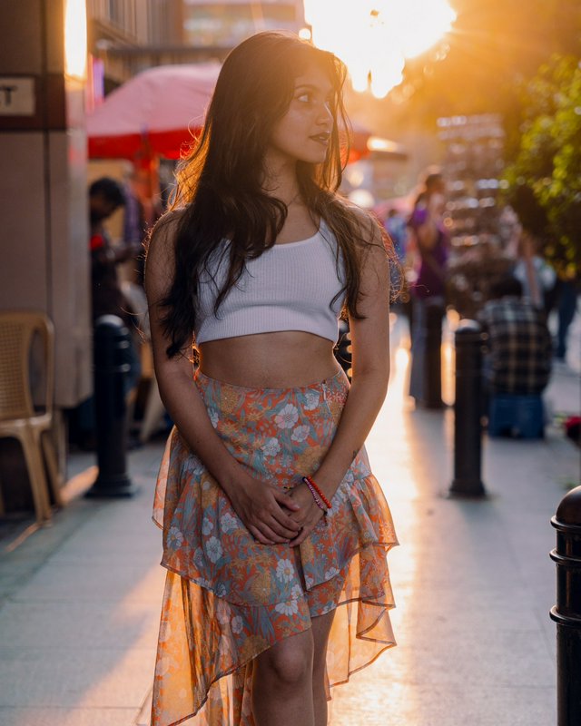 Young woman in white crop top and tiered floral skirt stands on a busy city sidewalk at golden ho...