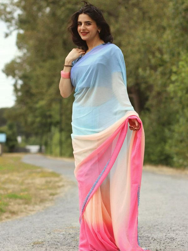 Smiling woman wearing a gradient pastel chiffon saree stands on an outdoor tree-lined gravel path