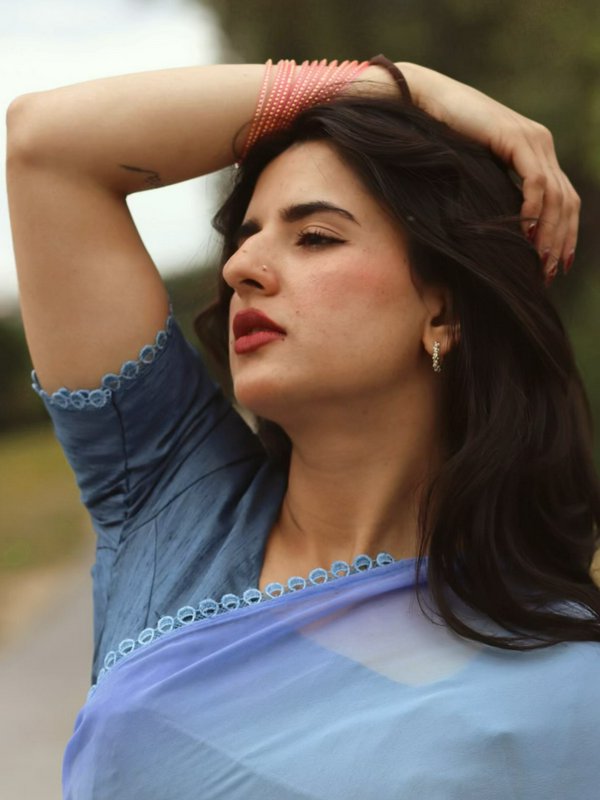 Closeup portrait of South Asian woman in light blue saree, arm raised over her head, soft outdoor...