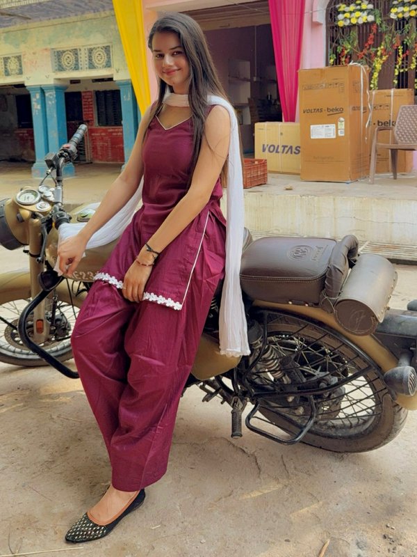 Smiling young woman in maroon salwar suit sits perched on a vintage golden motorcycle outside a r...