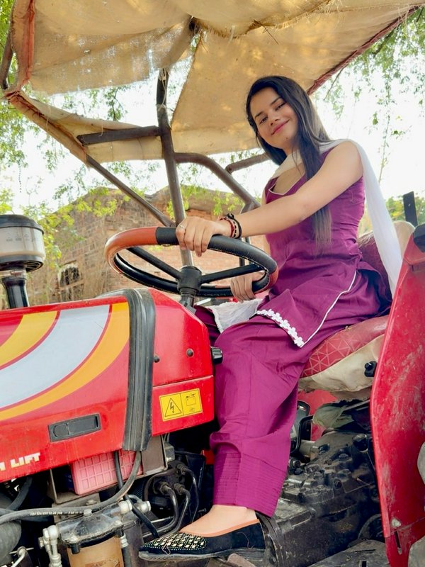 Young South Asian woman in a purple salwar suit smiles while holding the steering wheel of a red ...