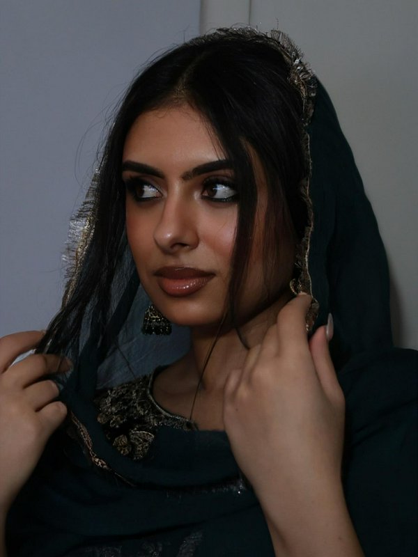 Closeup portrait of a young South Asian woman adjusting her gold-trimmed dark dupatta, gazing thoughtfully off to the side.