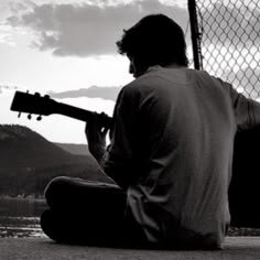 Black-and-white photo of a person sitting with a guitar by a chain-link fence near a lake at sunset.