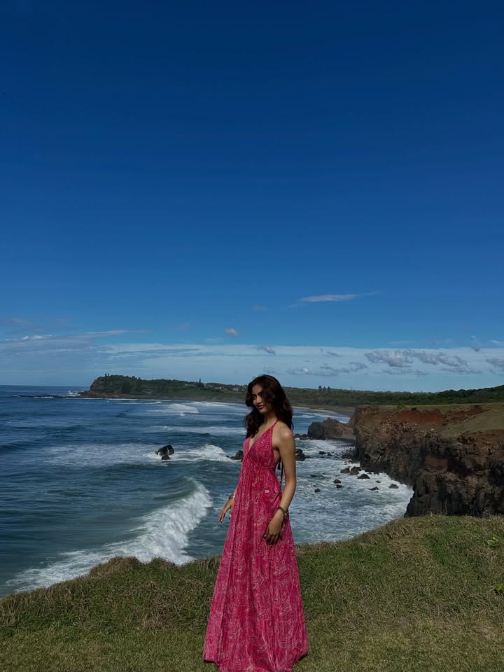 Woman in a pink dress standing on a grassy coastal cliff overlooking the ocean.