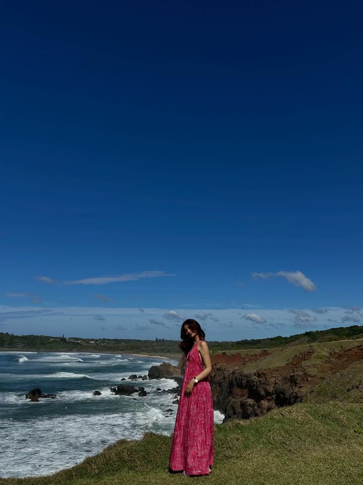 Woman in a pink maxi dress standing on a grassy cliff overlooking a rocky coastline and blue ocean