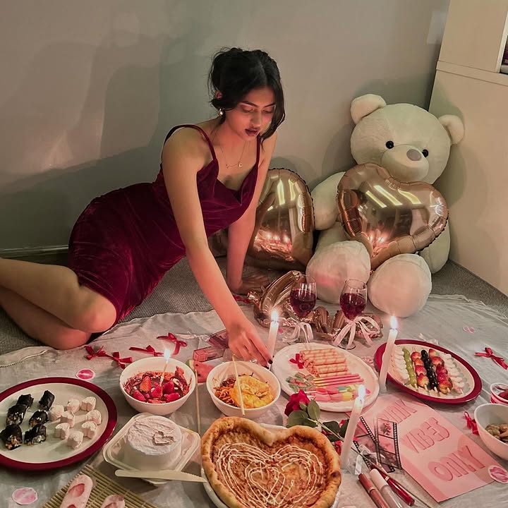 Woman in a burgundy dress arranging a heart-shaped pizza and sweets for a cozy birthday setup with a teddy bear balloon.