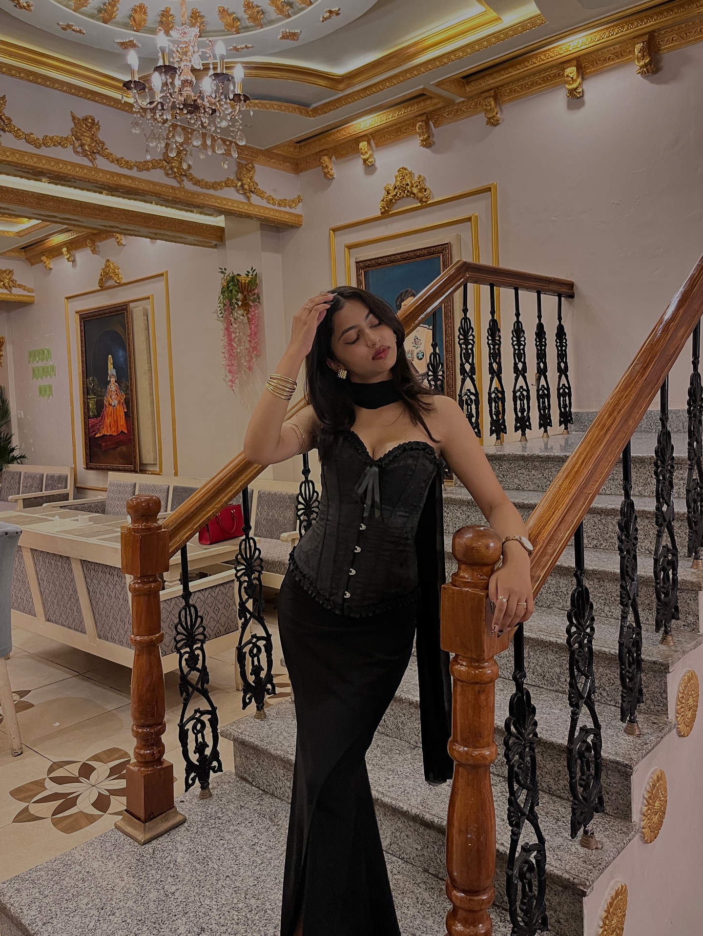 Woman in a black corset gown posing on an ornate staircase with gold detailing and chandelier.