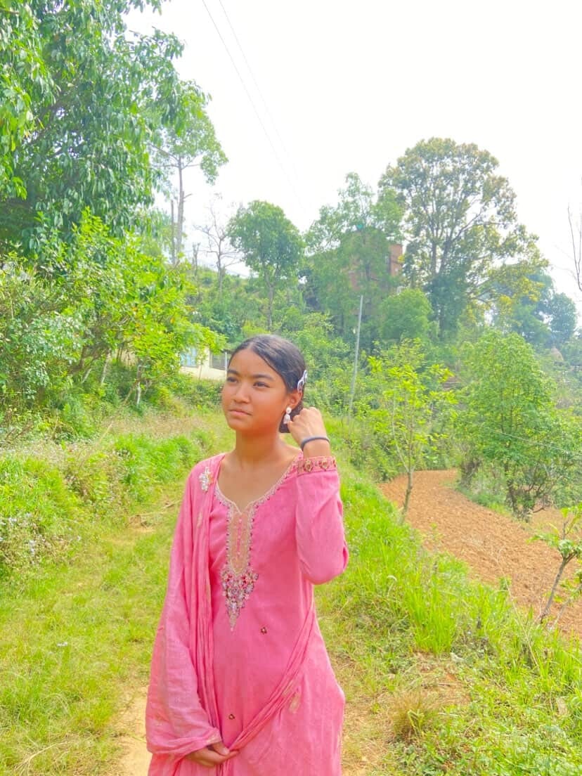 Young woman in a pink traditional kurti standing on a rural dirt path among green trees, adjusting her earring.