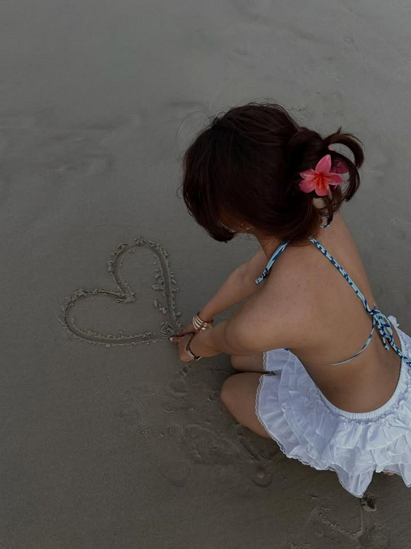 A woman in a white dress drawing a heart in the sand at the beach with a pink flower in her hair.