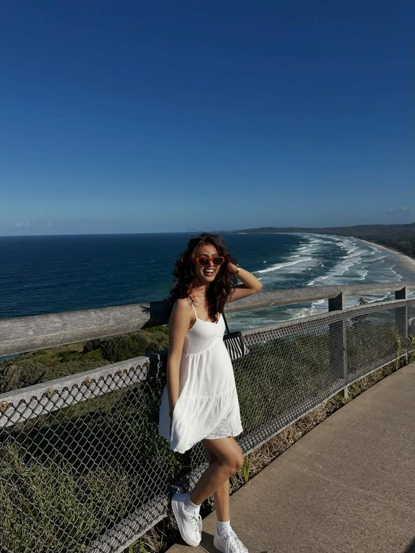 Woman in a white sundress and sneakers posing by a metal railing with a coastal ocean view.