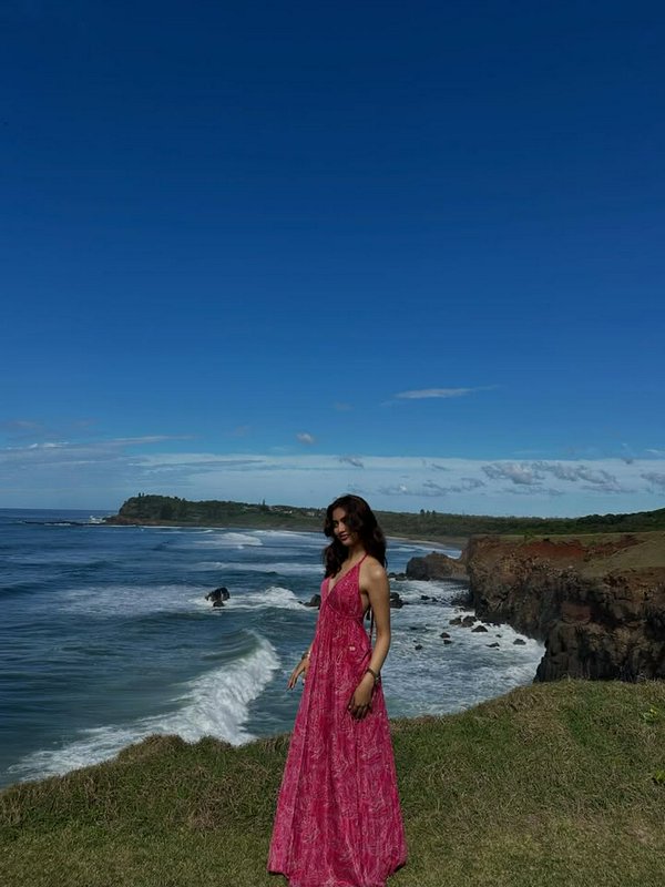 Woman in a pink dress standing on a grassy coastal cliff overlooking the ocean.