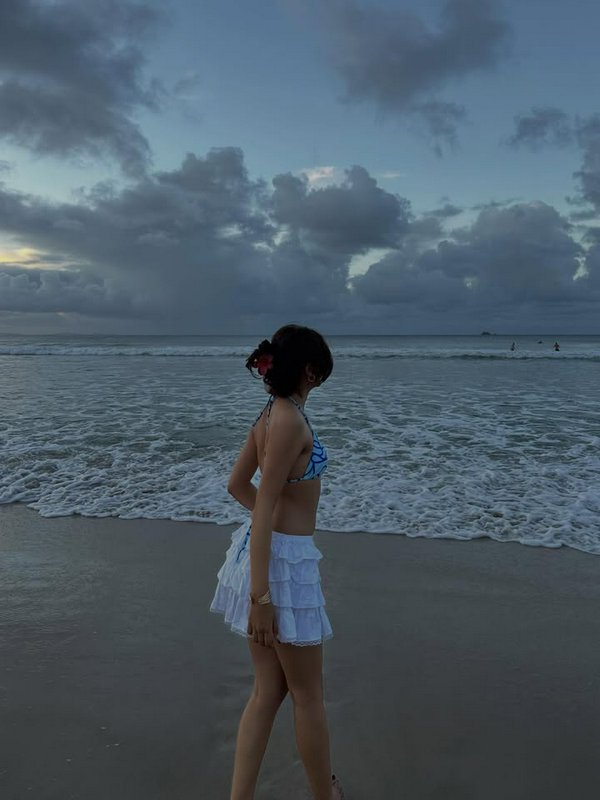 Woman in a blue bikini and white ruffled skirt standing on a sandy beach at dusk.