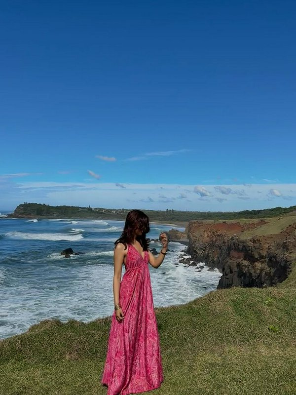 Woman in a pink dress standing on a grassy coastal cliff overlooking the ocean.