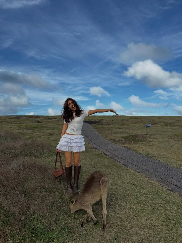 Woman in white top and layered skirt with knee-high boots posing beside a grazing deer on a grassy trail under a blue sky.