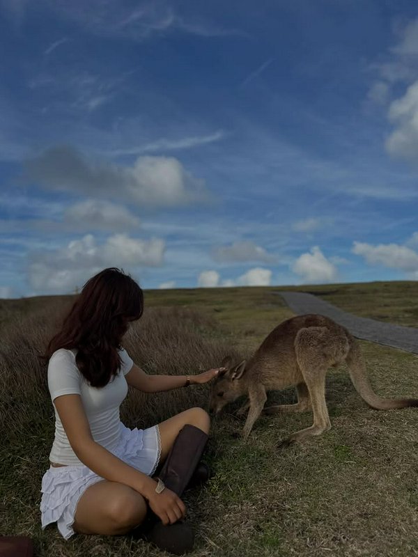 Woman in white sits on grass, gently petting a kangaroo in an open field under a blue sky.