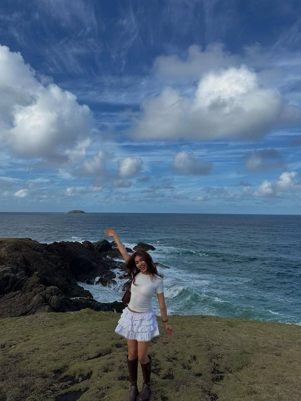 Woman in white top and ruffled skirt standing on rocky coastline, waving by the ocean under a blue sky.