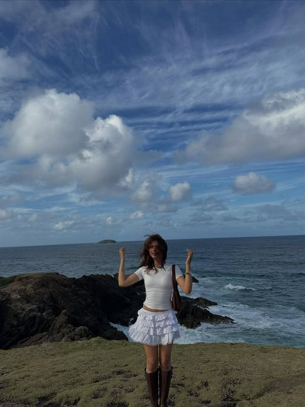 Woman in white top and ruffled skirt standing on coastal rocks by the sea under a blue sky.