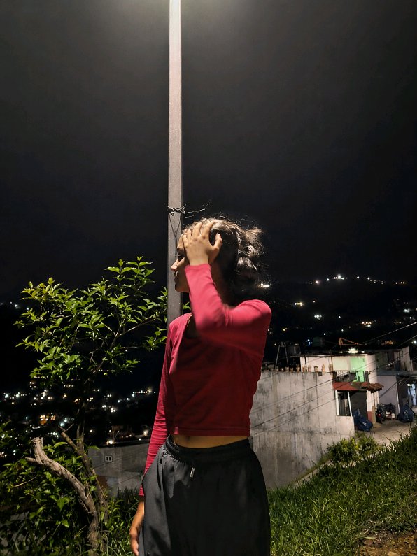 Young woman in a red long-sleeve crop top stands by a streetlight at night, overlooking a lit hil...
