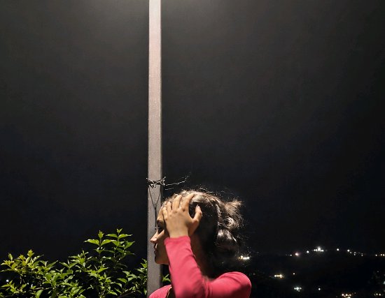 Young woman in a pink top holds her head on a night balcony, overlooking distant city lights with...