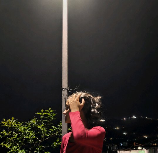 Profile of a woman in a red long-sleeve top with her hand on her head, standing by a pole at nigh...